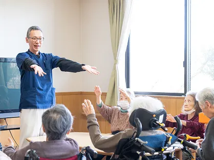 介護施設のレクリエーションの様子。スタッフが両手を広げながら笑顔で高齢の方に呼びかけ、車椅子に座った複数の入居者が手を挙げて楽しそうに参加している。